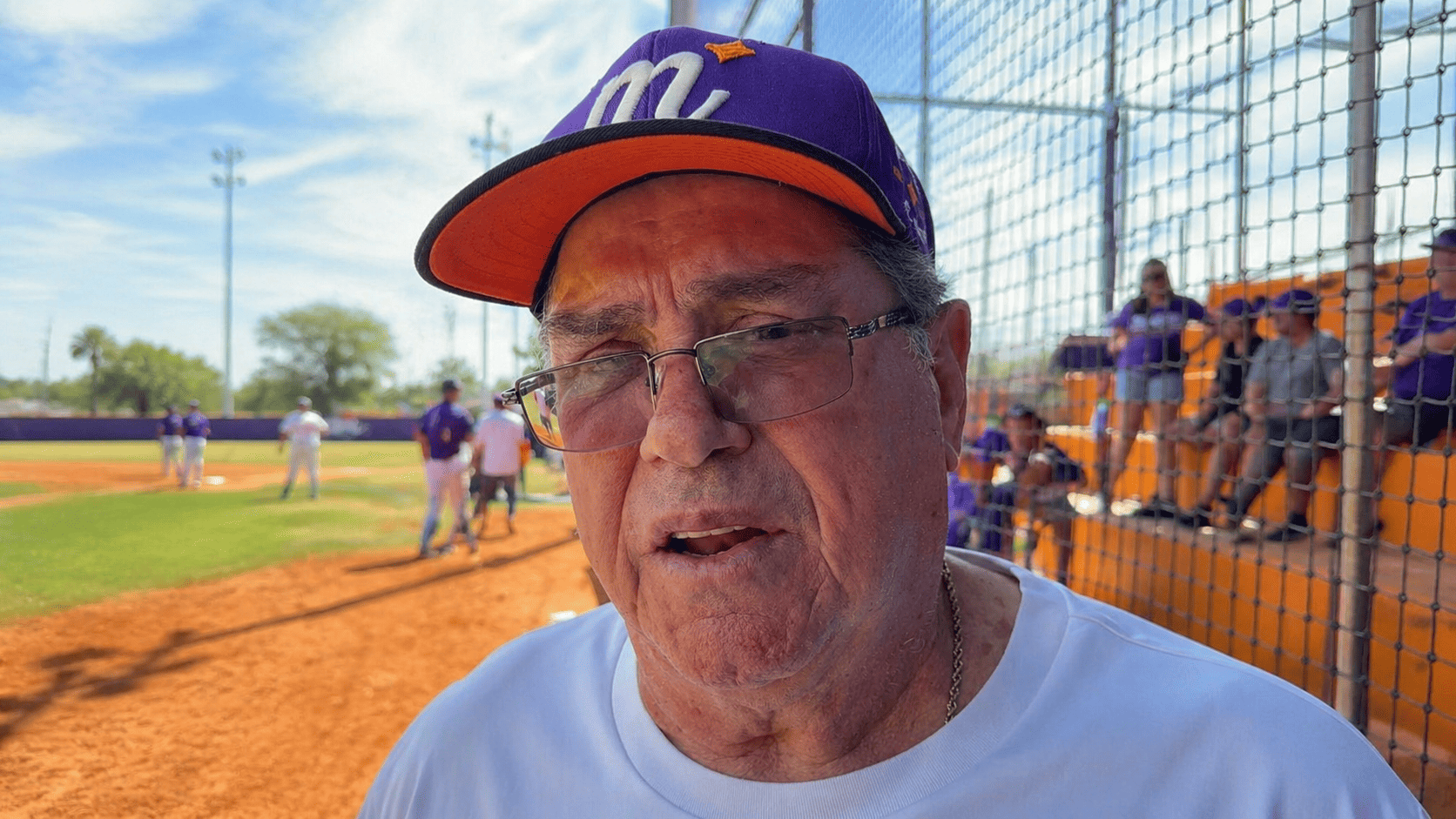 Mario Mendoza aparece en primer plano durante una entrevista en un campo de beisbol, con gorra morada y naranja, lentes y graderío al fondo.