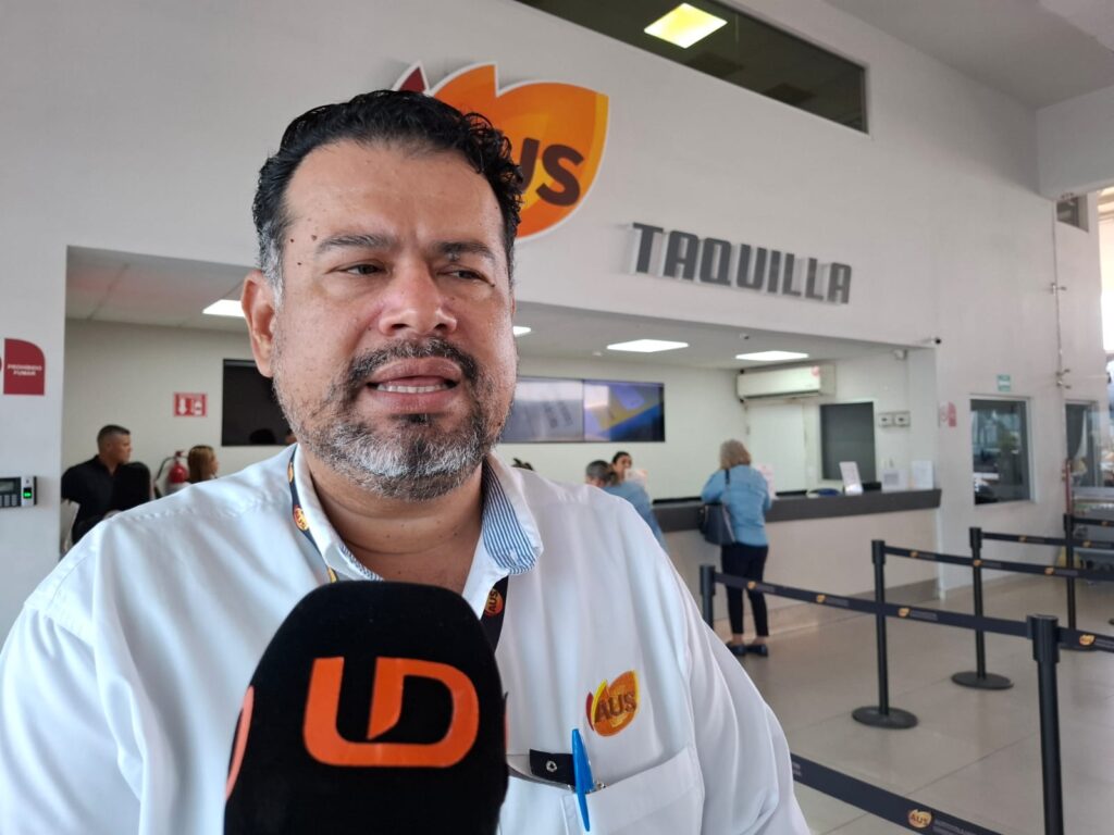 Persona viendo hacia el frente con un micrófono negro con las letras LD en color naranja, en una central de autobuses