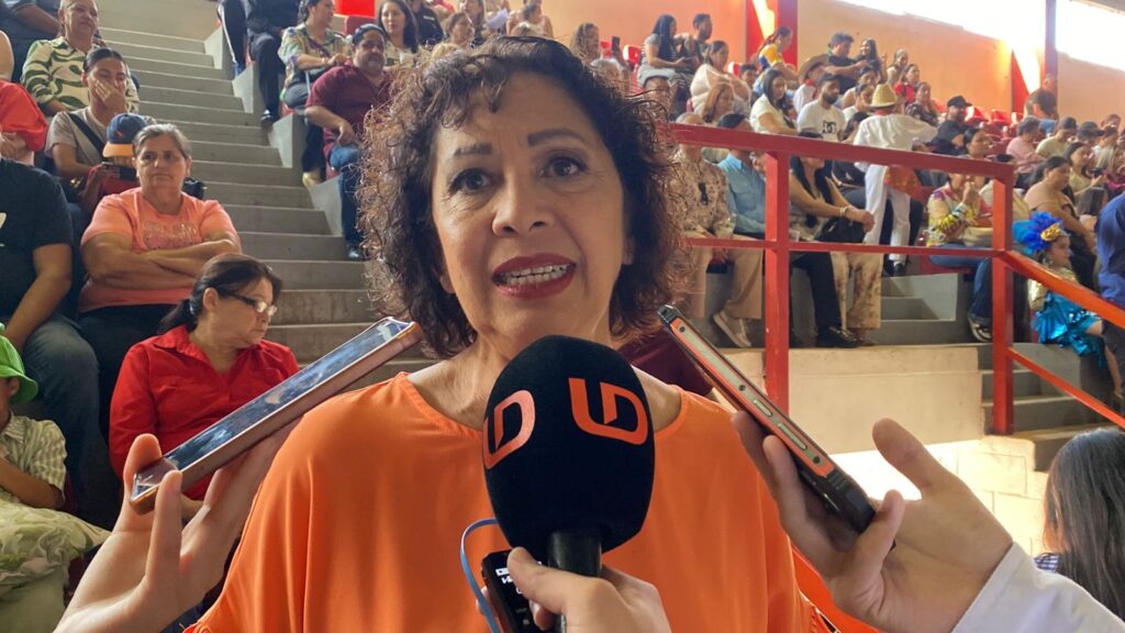 mujer hablando hacia el frente con un micrófono y al fondo personas en un auditorio