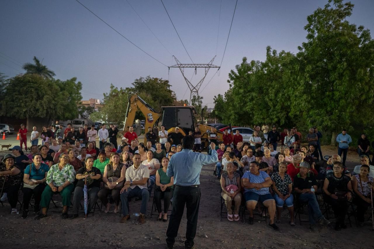 Personas sentadas en sillas a la intemperie y detrás de ellos maquinaria de construcción, mientras el alcalde habla al frente por micrófono