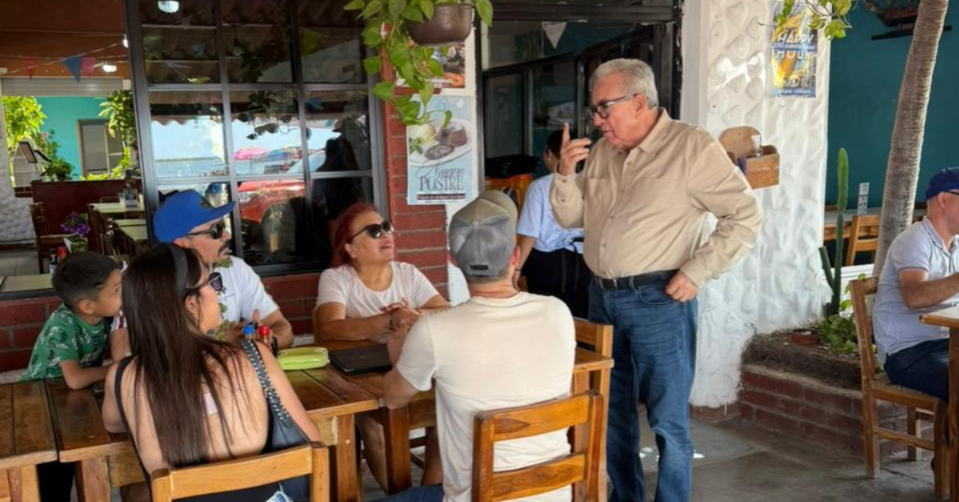 Cuatro personas sentadas en una mesa junto a el gobernador parado platicando