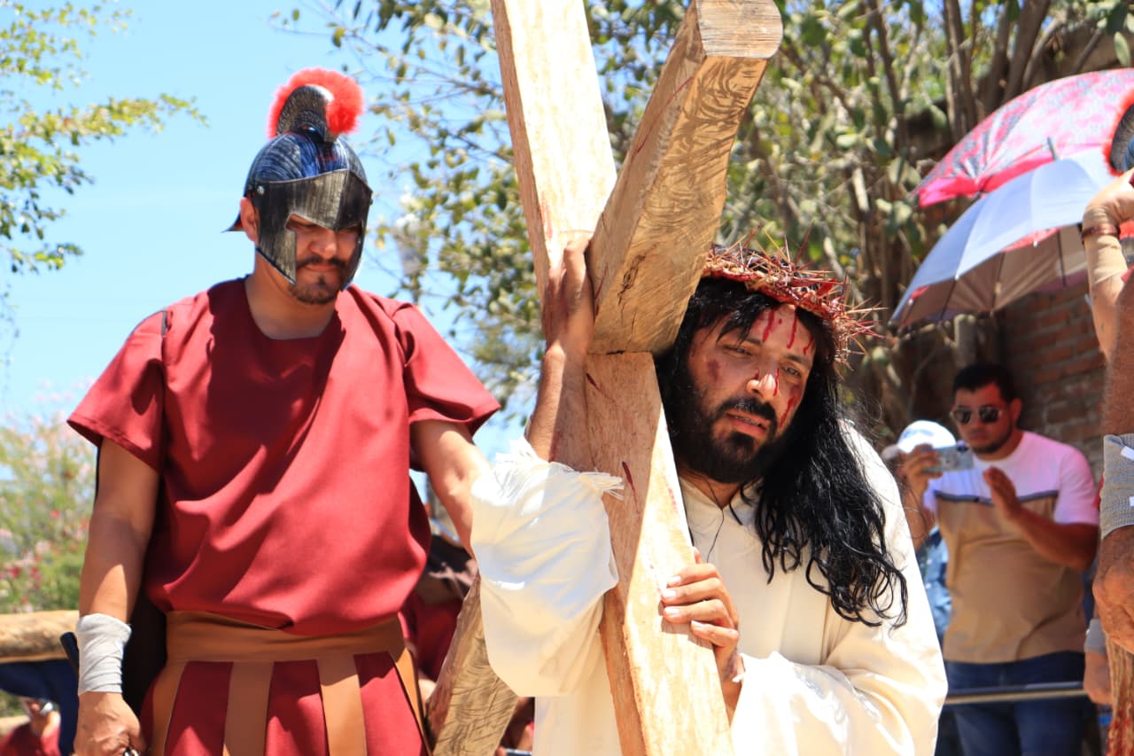 Persona cargando una cruz de madera con una corona de espinas y sangre en la cara, junto a una persona con casco negro con rojo