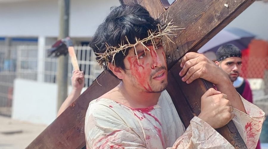 Un hombre con una corona de espeinas en la cabeza con manchas rojas en la cara y cargando una cruz de madera