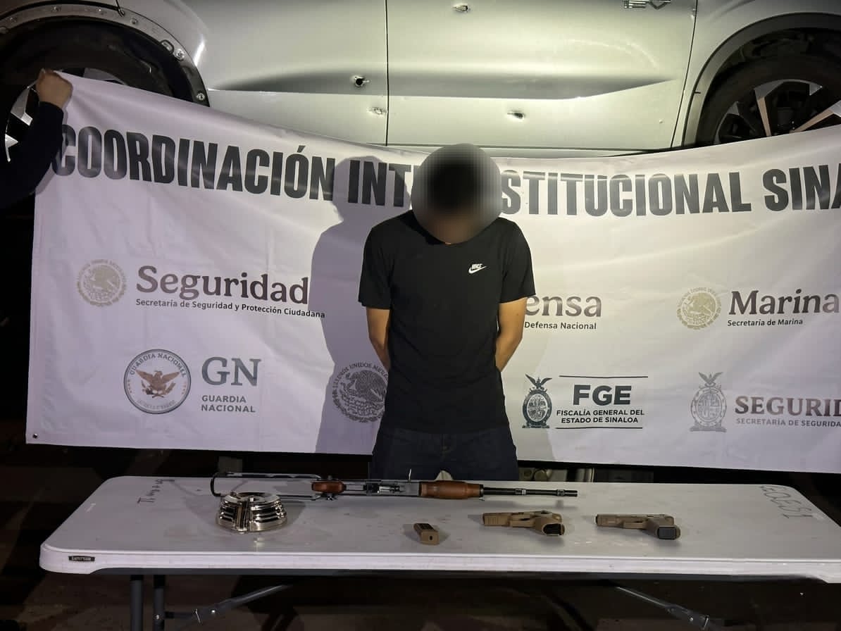 persona con el rostro cubierto y la cabeza agachado y en una mesa armas y objetos asegurados, al fondo una lona con logos de corporaciones policiacas