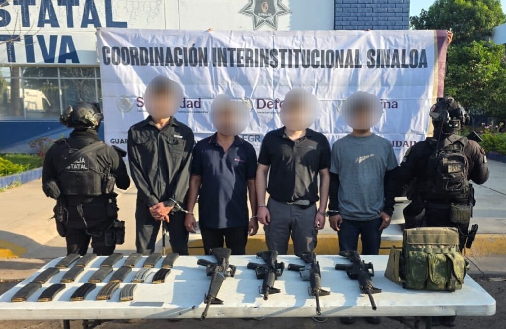 cuatro personas con el rostro cubierto y dos agentes policiacos a las orillas, frente a ellos una mesa con armas, equipo táctico y cargadores y al fondo una lona con logos de corporaciones de seguridad