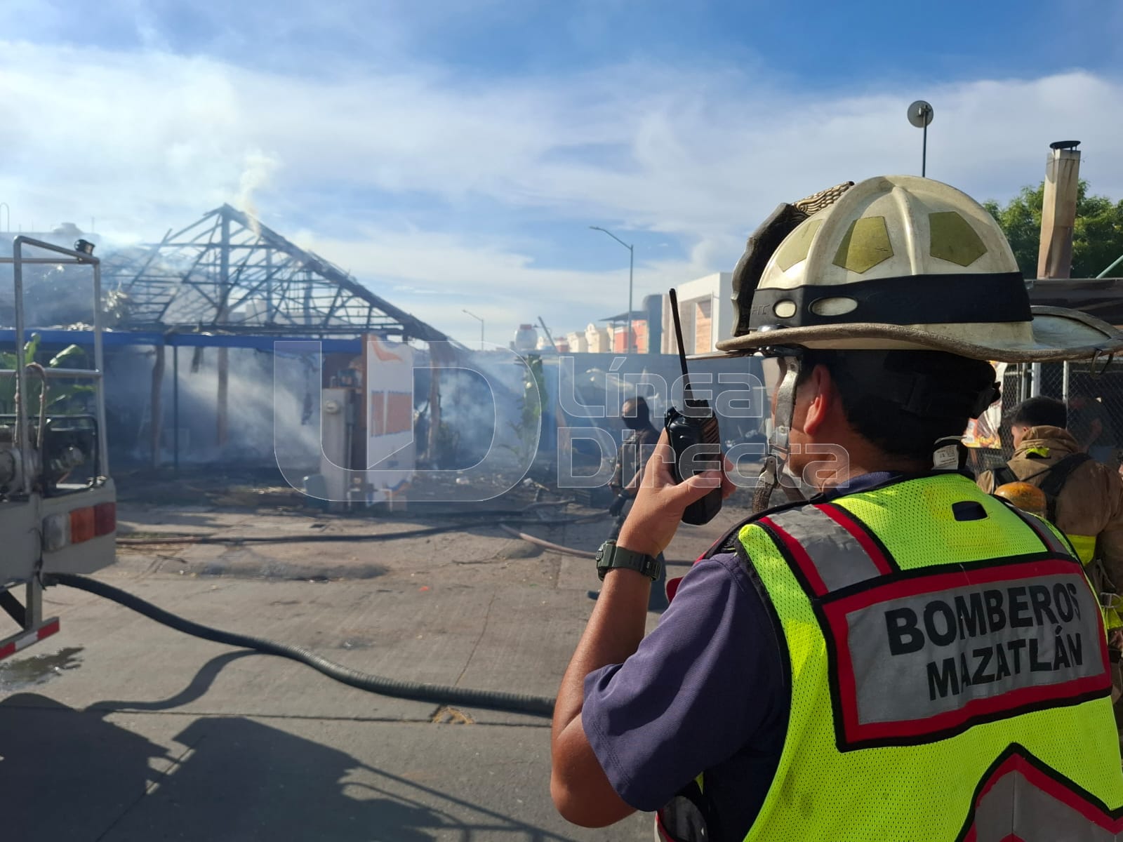 Elemento del cuerpo de Bomberos Mazatlán con equipo y un radio frente a una palapa entre humo