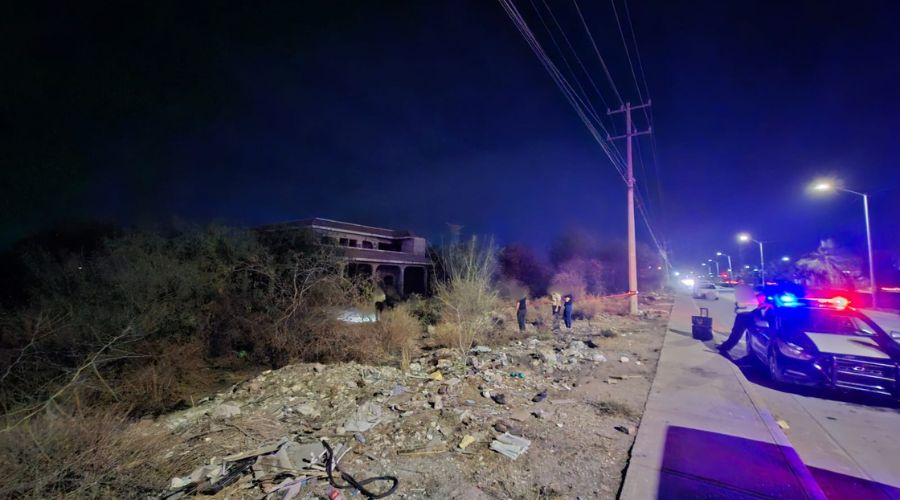 Unidad policiaca estacionada cerca de una casa abandonada entre matorrales y personas