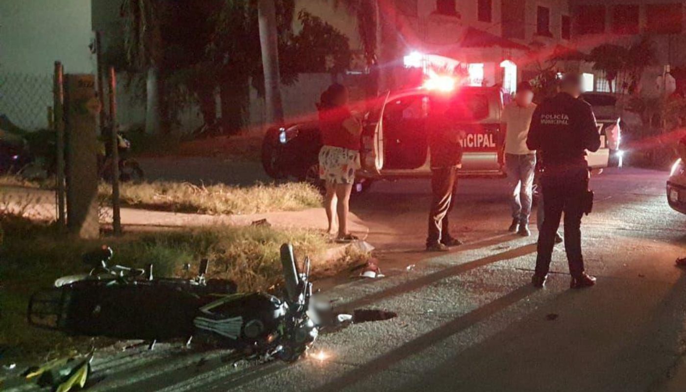 Motocicleta tirada en una calle junto a unas personas y agentes policiacos, al fondo una patrulla con las luces encendidas