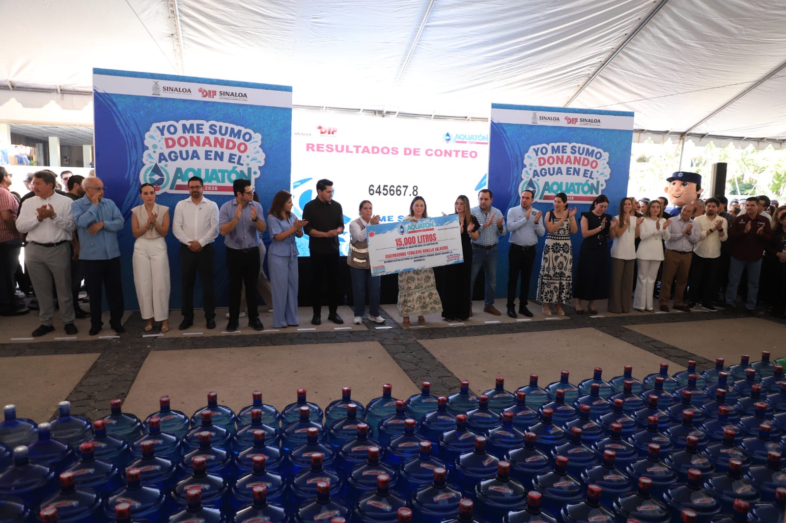 evento público de donación de agua, Hay una gran cantidad de garrafones de agua purificada, Una fila de personas, que parecen ser funcionarios públicos, organizadores y asistentes, están de pie frente a los garrafones aplaudiendo de manera celebratoria.