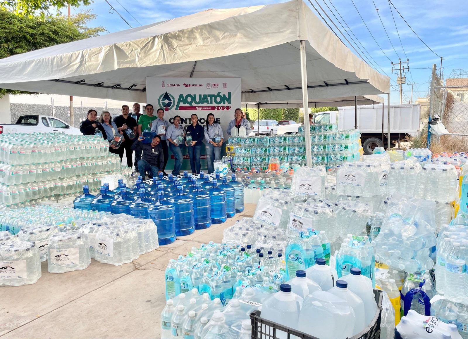 Centro de acopio de agua, con numerosos paquetes de agua embotellada y al fondo personas posando para la foto bajo una carpa