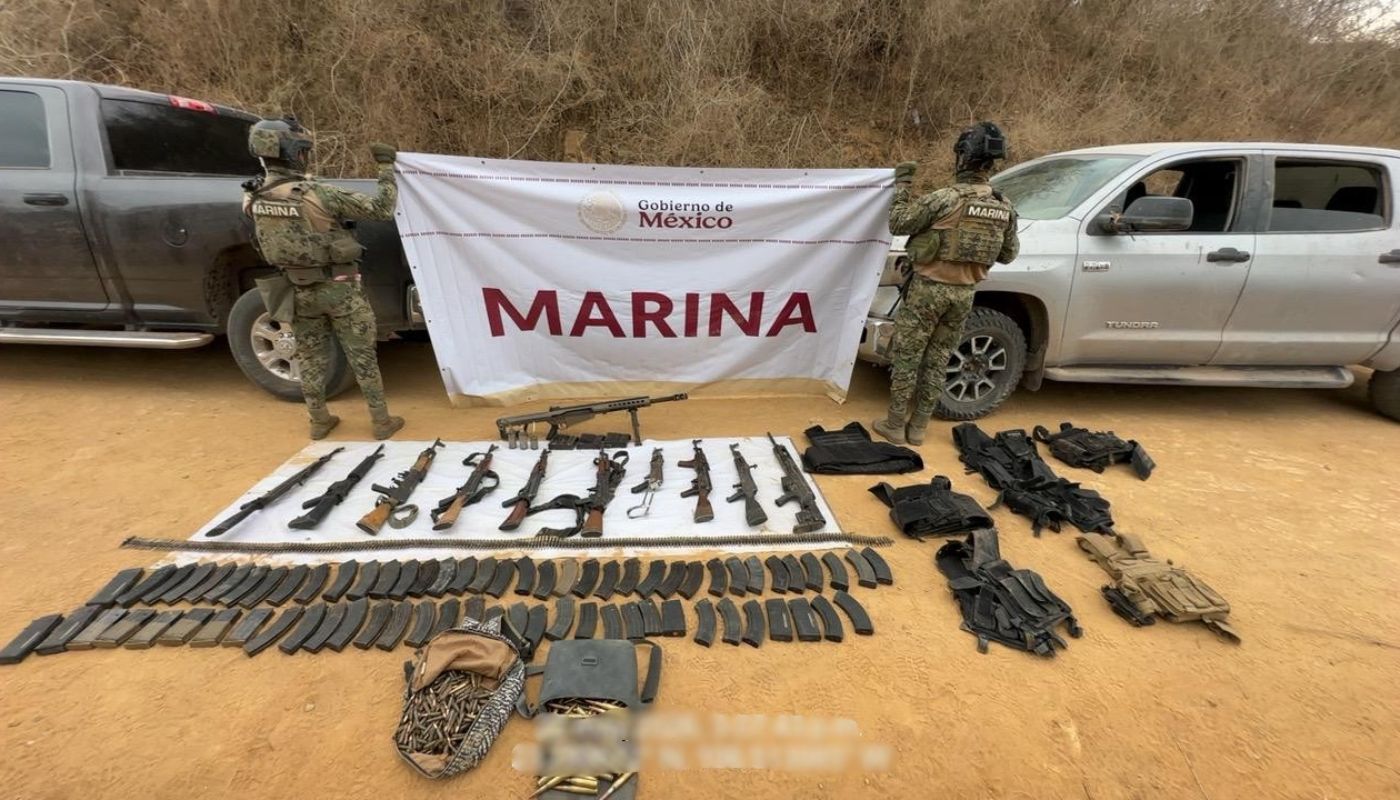 Elementos de la marina de espaldas sosteniendo una pancarta, en el suelo armas y artefactos confiscados, a los costados unidades aseguradas