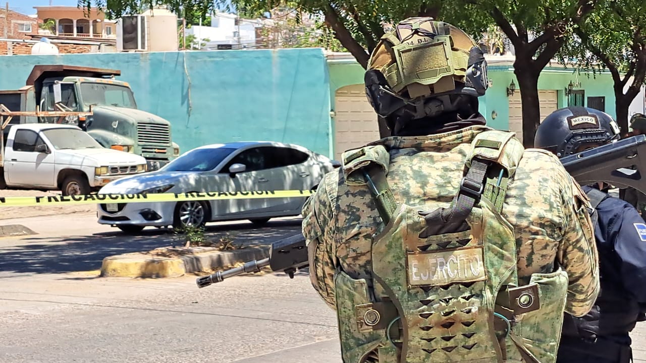 Elemento militar de espaldas, al fondo un auto blanco detrás de una cinta amarilla acordonando una área en plena calle