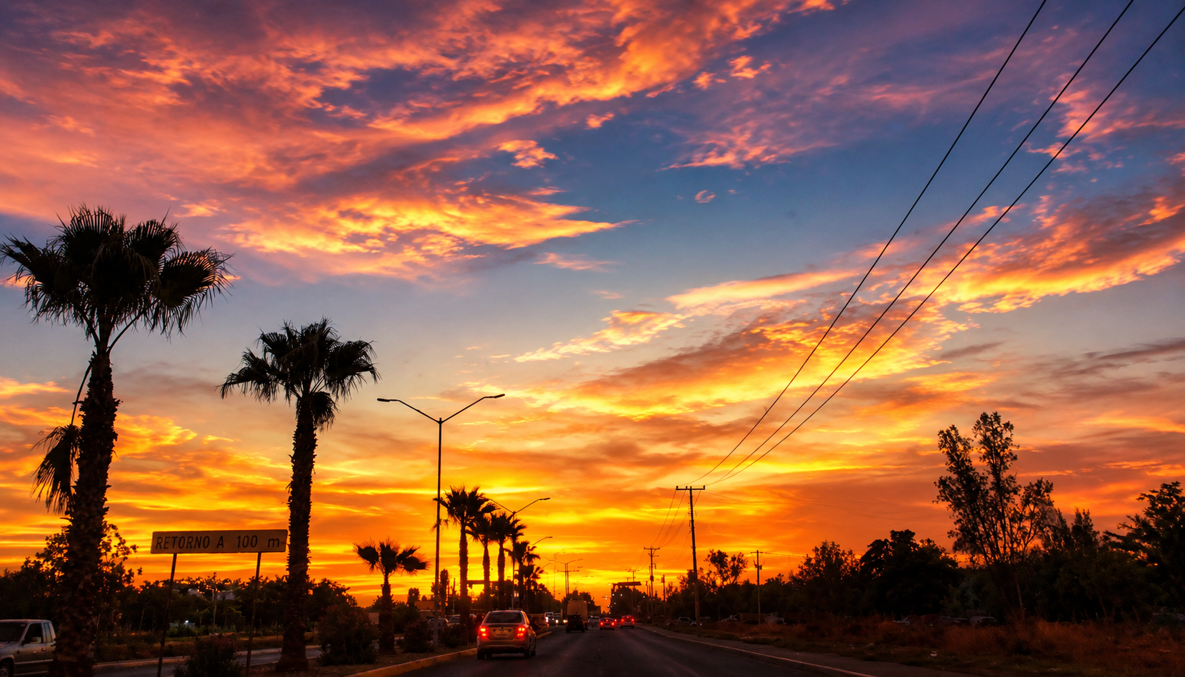 Atardecer con cielo naranja, rosa y violeta sobre una avenida con palmeras, vehículos y líneas eléctricas al fondo.