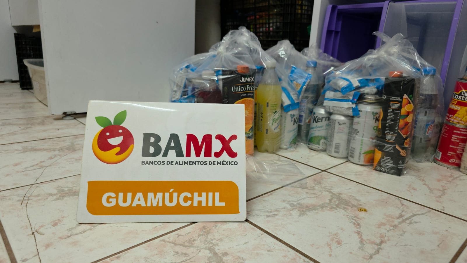 Cartel pequeño de Banco de alimentos de México con bolsas con despensas detrás en el piso