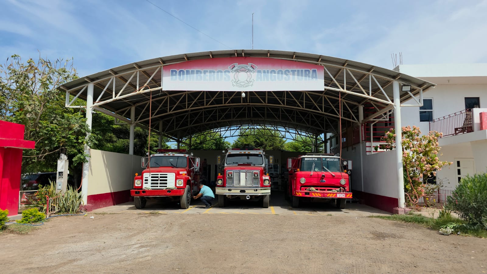 Vista frontal de la estación de Bomberos de Angostura con tres camiones de emergencia estacionados bajo una estructura metálica durante el día.