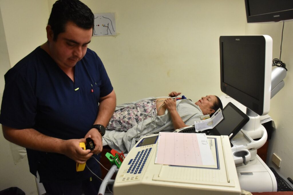 Persona acostada en una camilla junto a un médico con aparato de cardiología