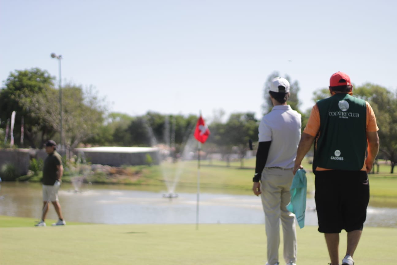 Dos golfistas y un caddie observan el green junto a un lago con fuentes en un campo de golf soleado, mientras otro jugador camina al fondo.