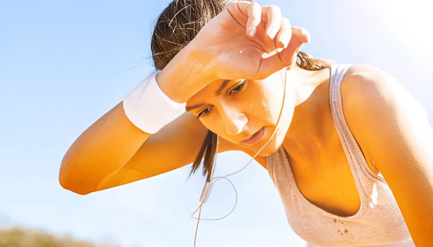 una mujer joven al aire libre, experimentando lo que parece ser un intenso calor, iene el brazo levantado apoyando la mano sobre su frente