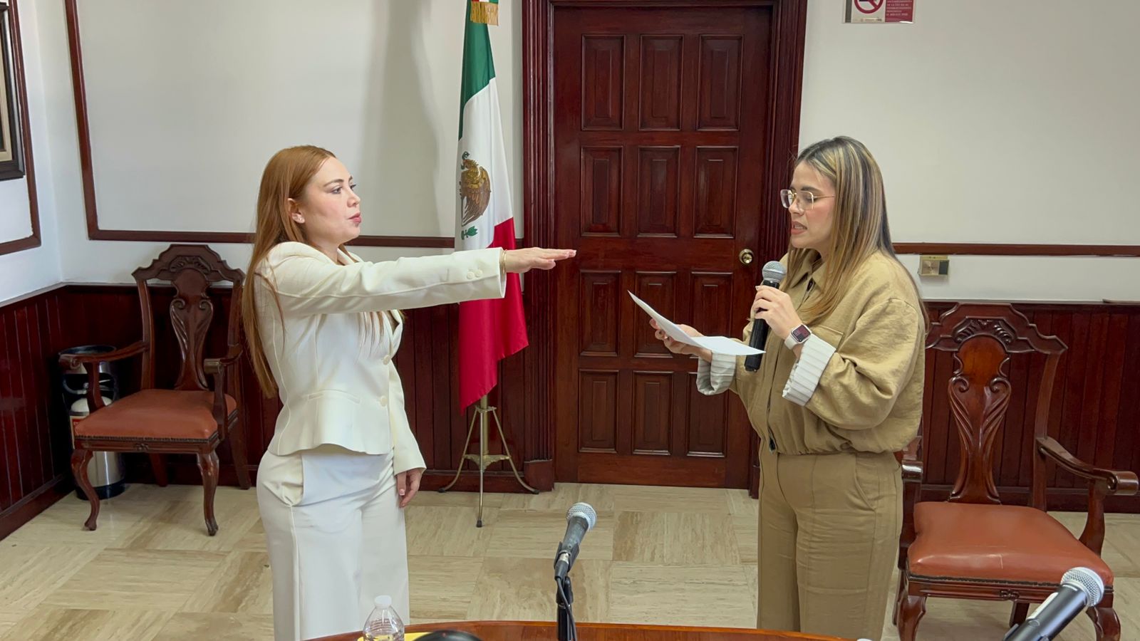 Una persona levantando la mano en señal de protesta frente a otra persona, en una oficina con una bandera de México