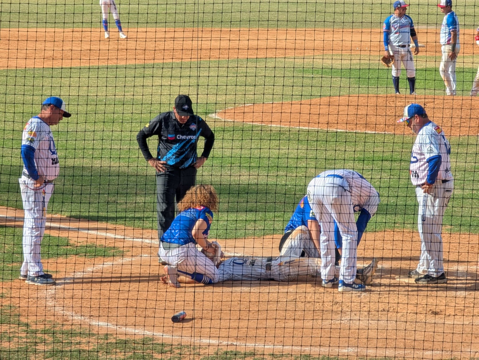 Jugadores de beisbol en terreno de juego y uno de ellos tendido en el suelo siendo atenido