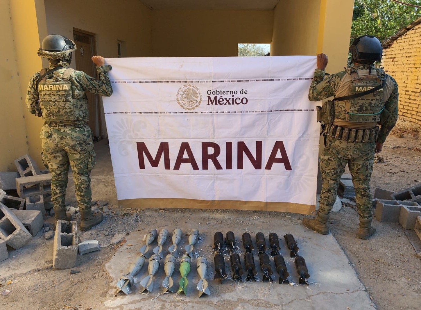 Militares con uniforme verde de espaldas sosteniendo lona de la Marina y artefactos decomisados en el suelo
