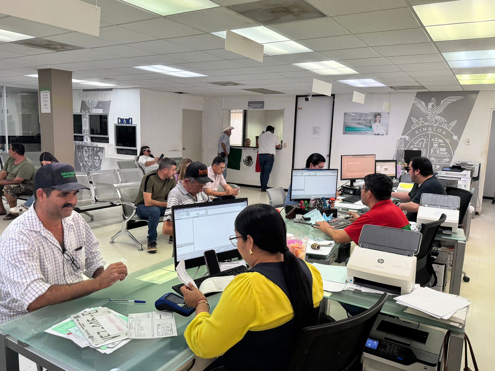 Personas realizan trámites y esperan turno en una oficina gubernamental de Guamúchil, mientras empleados atienden servicios administrativos y de licencias.