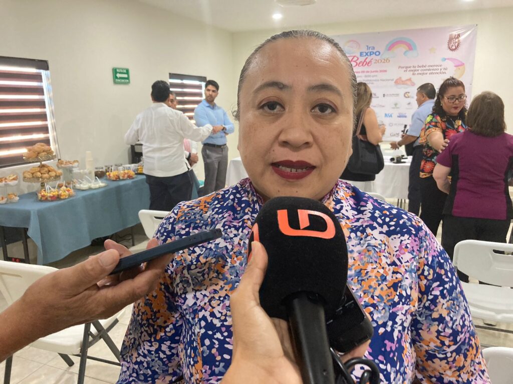 Mujer hablando con dos micrófonos frente a ella y al fondo más personas de pie