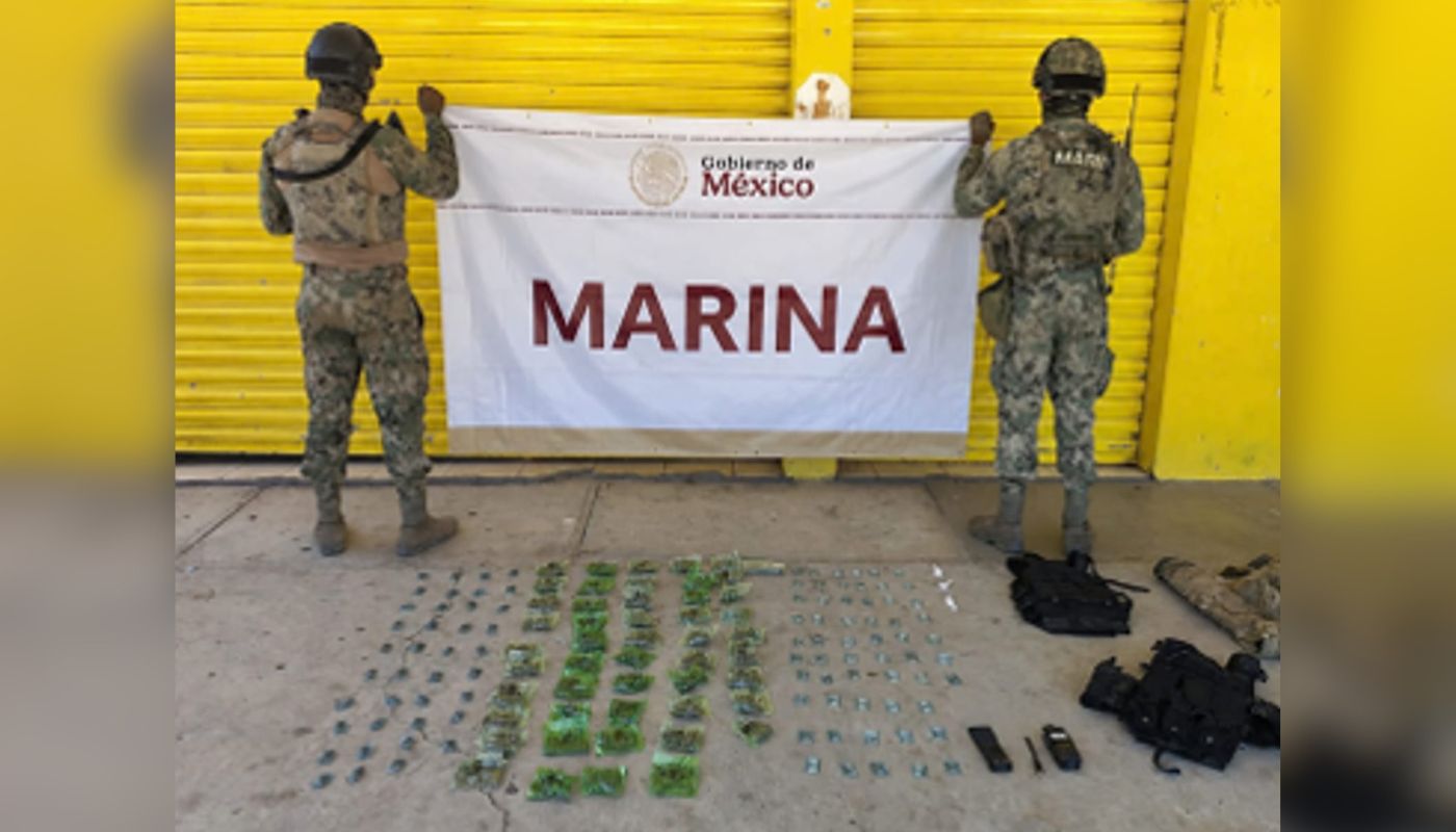 Dos elementos militares sosteniendo una lona con la leyenda MARINA y en la parte inferior otra lona con artefactos explosivos