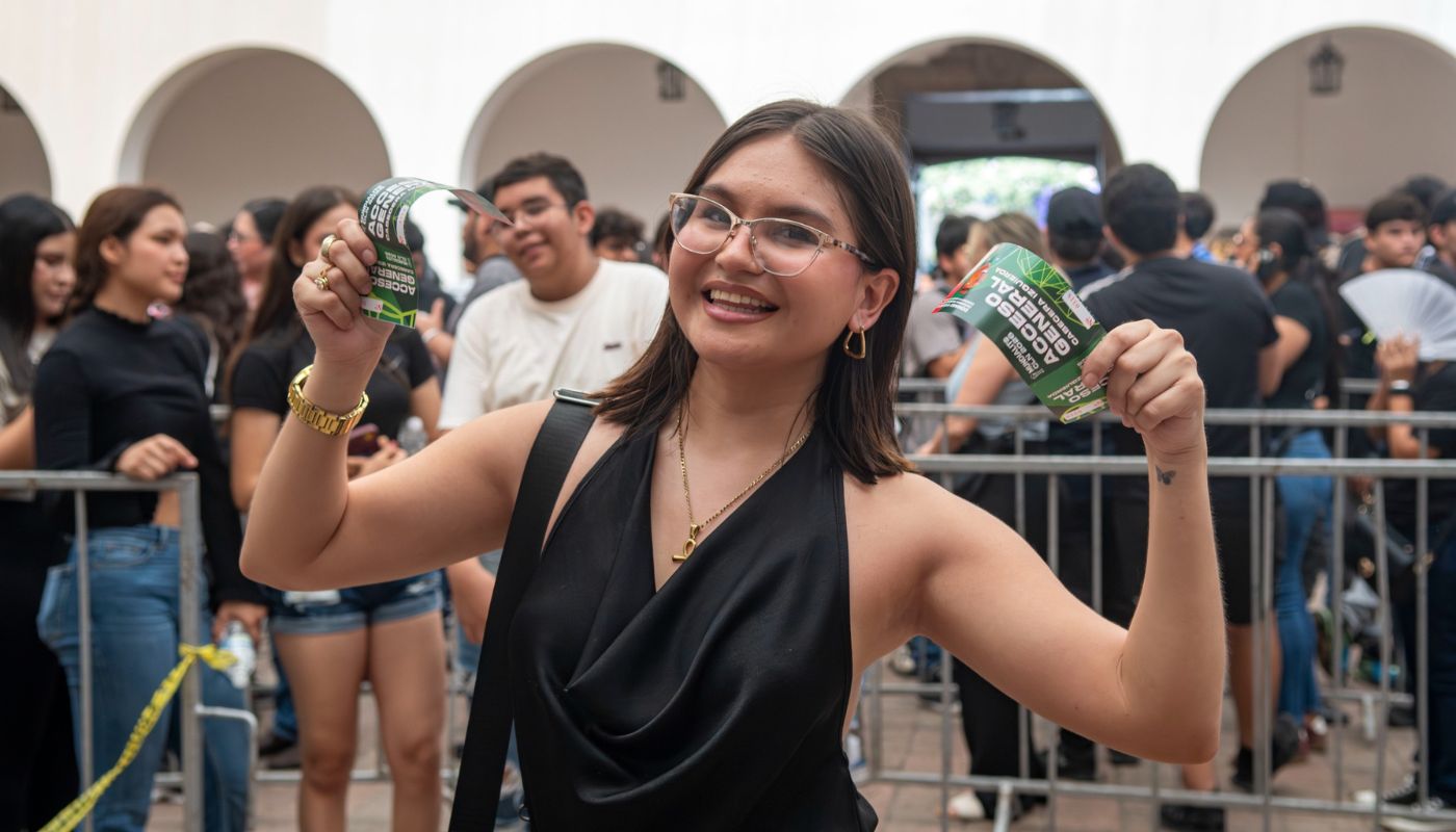 Mujer sosteniendo boletos en sus manos y al fondo personas haciendo fila