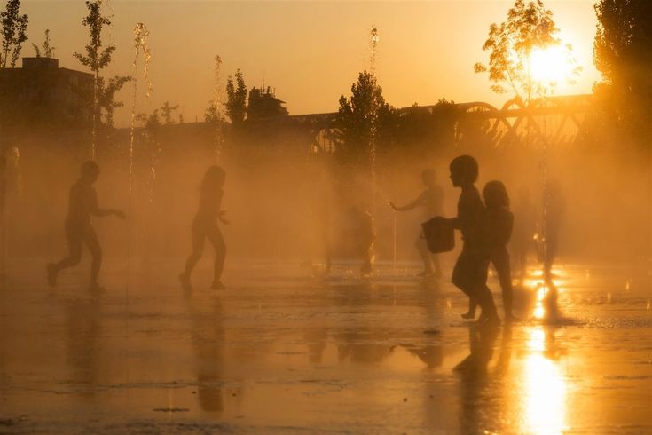 Niños jugando entre chorros de agua que surgen desde el suelo y un sol fuerte en el cielo naranja