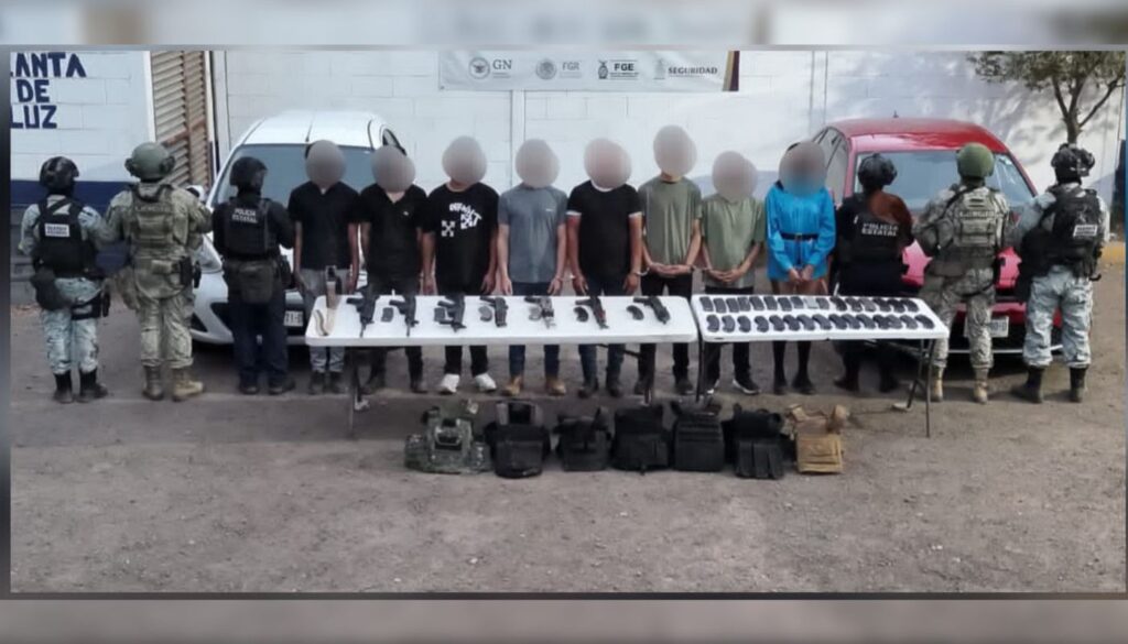 Agentes policiacos de espaldas y entre ellos 8 personas con el rostro cubierto y en una mesa armas y objetos