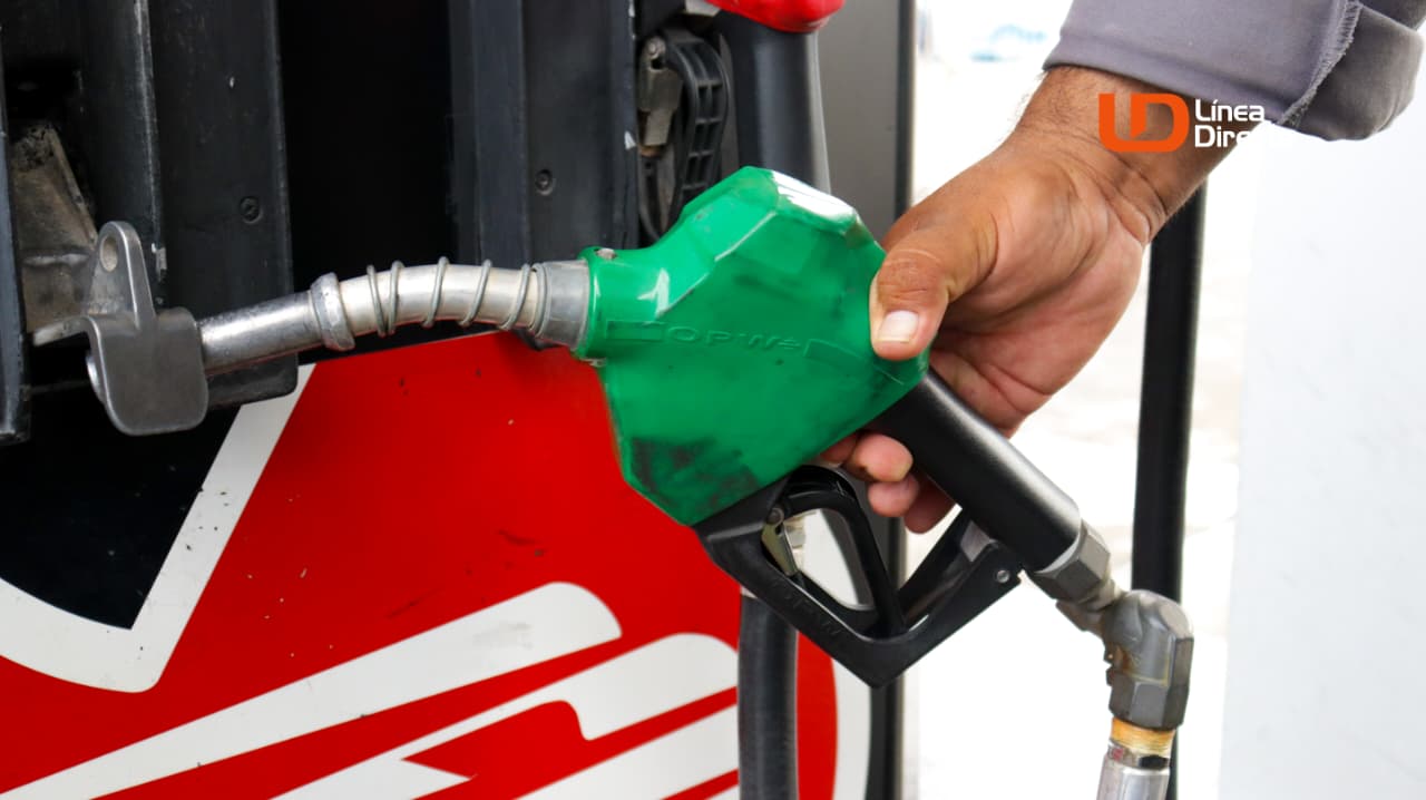 Mano sosteniendo una manguera dispensadora de gasolina de color verde y al fondo parte de una bomba de gasolina