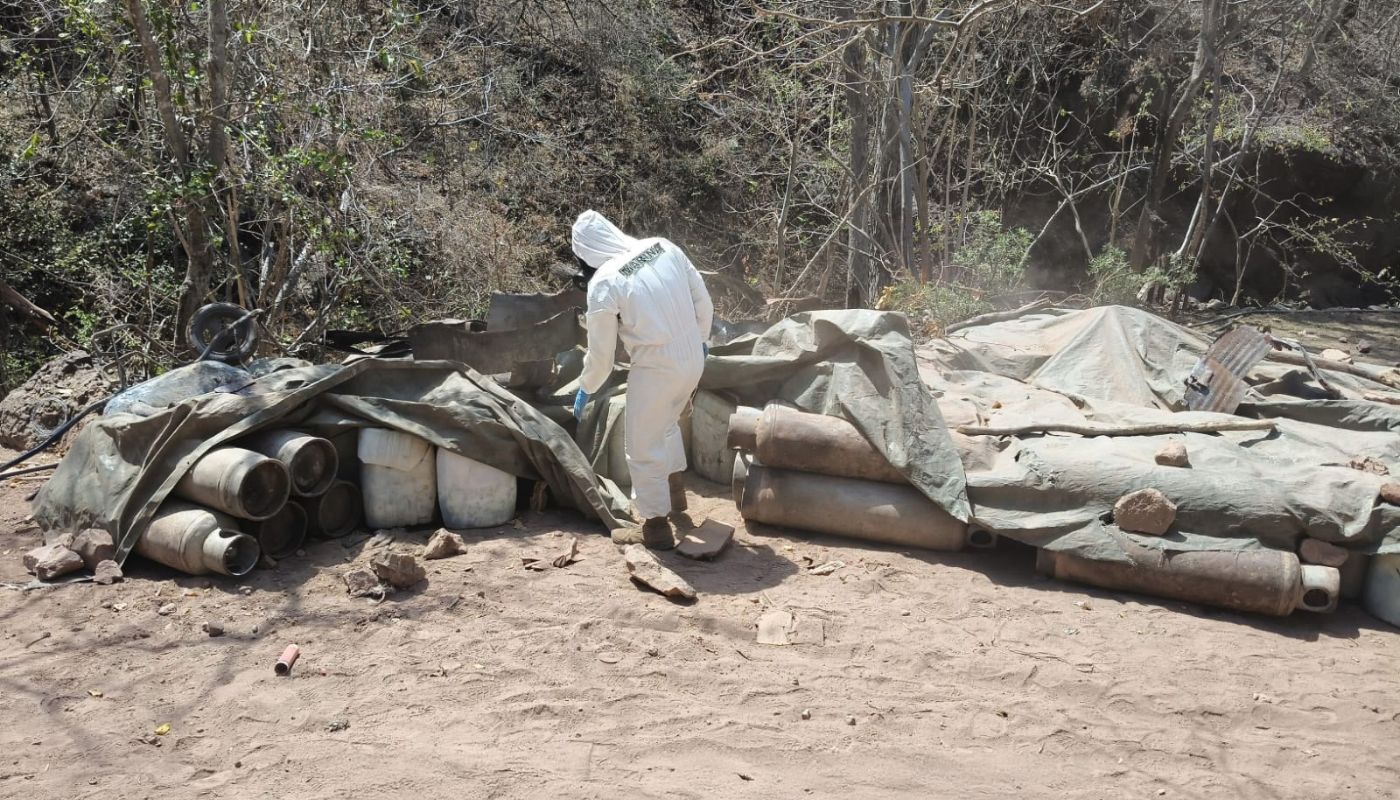 Agente de Fuerzas Armadas con un traje especial de color blanco entre objetos de un laboratorio clandestino en una zona tipo boscosa
