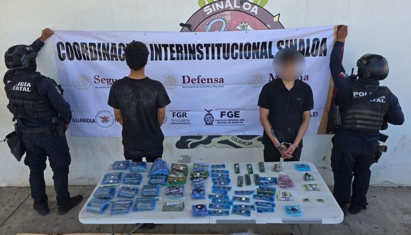 Elementos policiacos de espaldas sosteniendo una lona, al centro dos personas, una de espadas y otra de frente con las manos esposadas y el rostro cubierto, en la parte inferior una mesa con objetos/droga