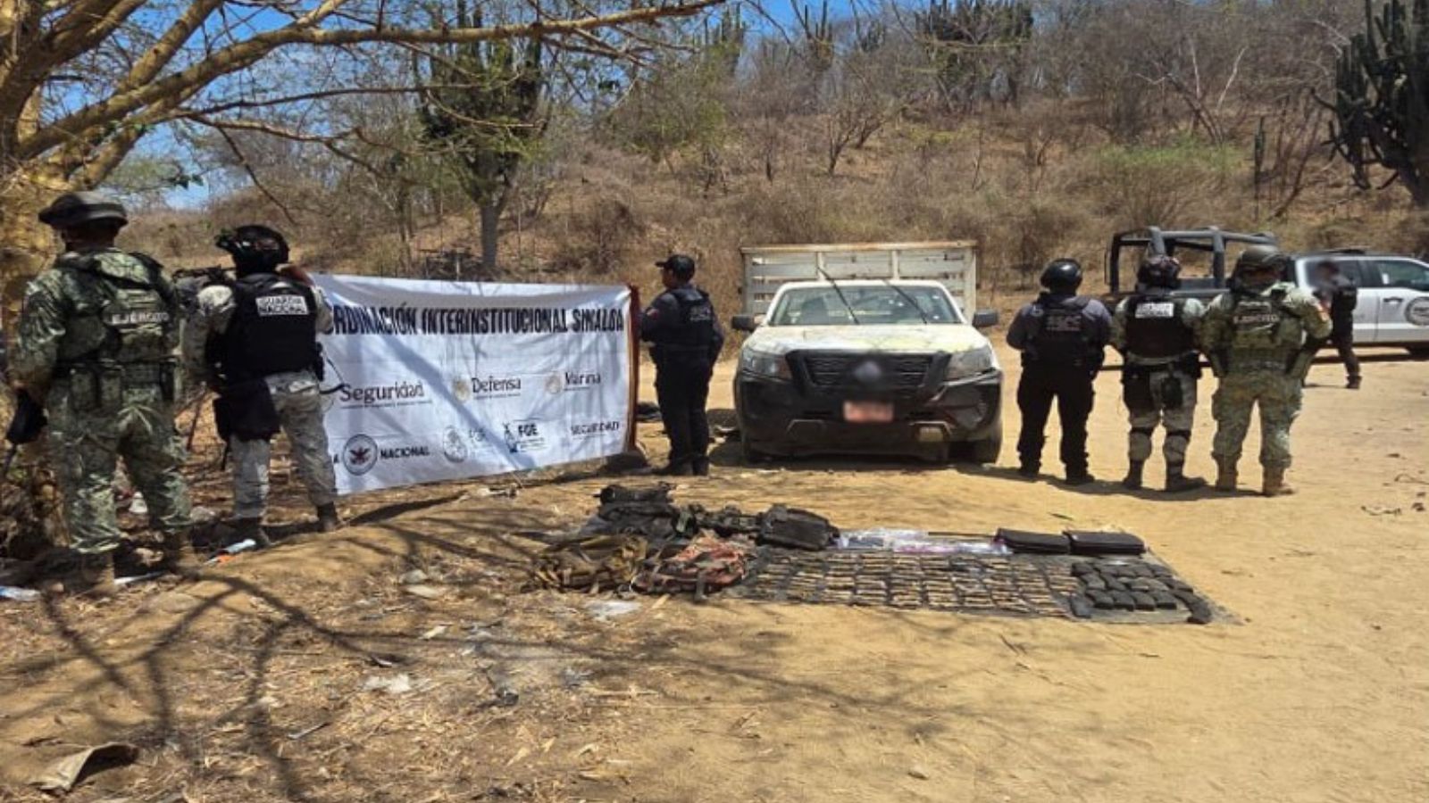 Elementos policiacos sosteniendo una lona con logos de corporaciones de seguridad junto a una camioneta de color blanco, en la parte inferior, una lona con