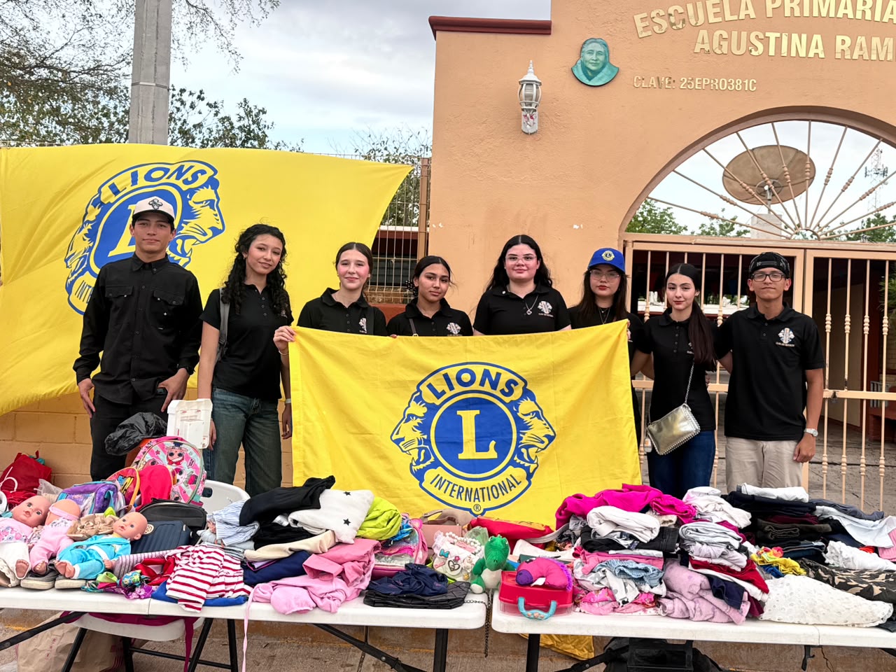 Jóvenes sosteniendo una lona/banner junto a una mesa con ropa y juguetes, afuera de una escuela