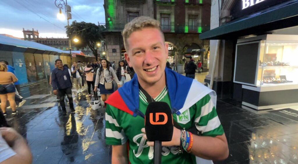 Daniel Hruby, influencer checo, con un jersey deportivo y una bandera en su espalda, en una calle y al fondo muchas personas