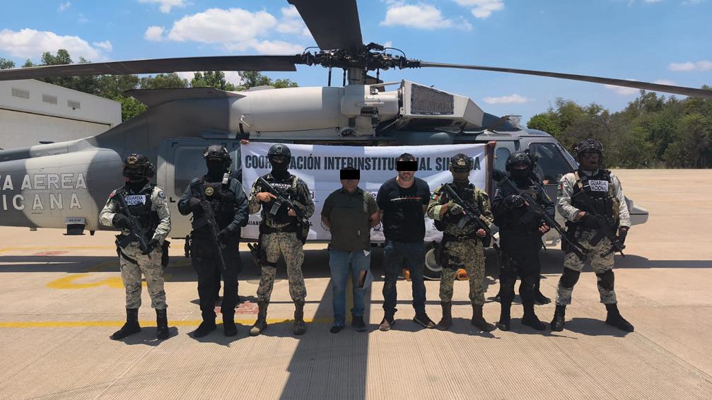 Agentes policiacos con armas y al centro dos civiles con el rostro cubierto y las manos esposadas, al fondo un helicoptero