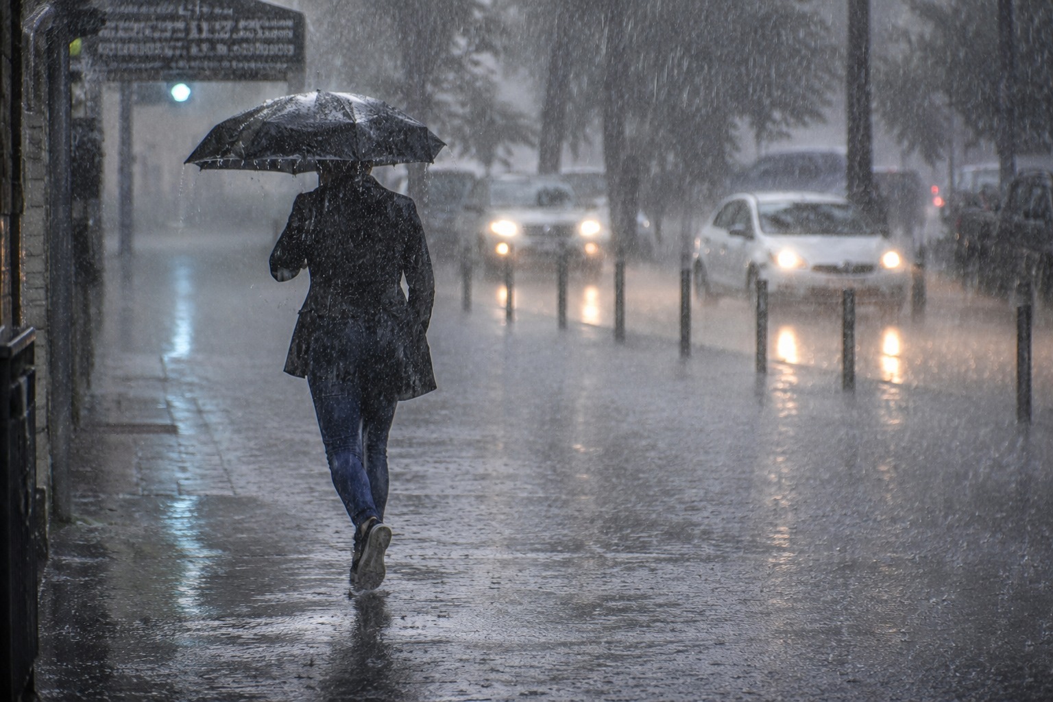 persona con un paraguas caminando junto a una calle con carros circulando con las luces encendidas en medio de la lluvia