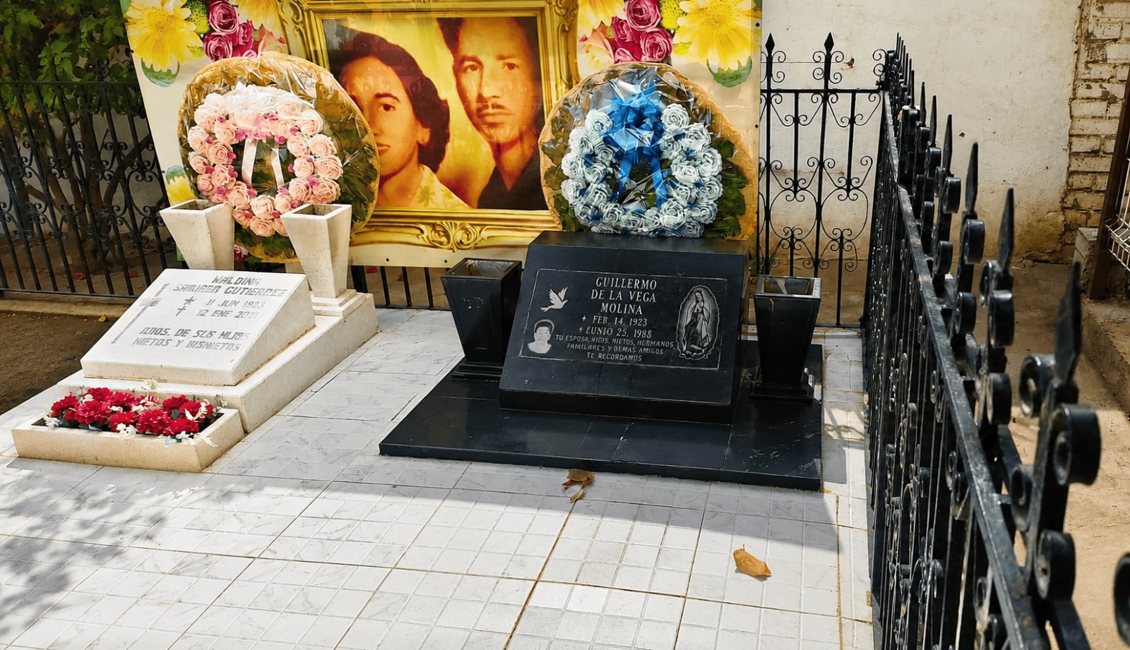 Tumbas decoradas con arreglos florales en un panteón, donde familiares visitan y honran la memoria de sus padres fallecidos mediante ofrendas y momentos de reflexión.