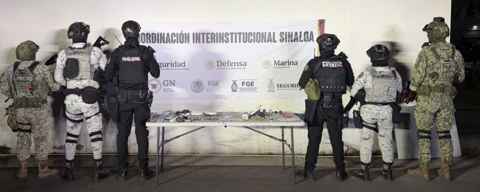 Cuatro elementos de seguridad con una mesa con armas y drogas y una lona de seguridad