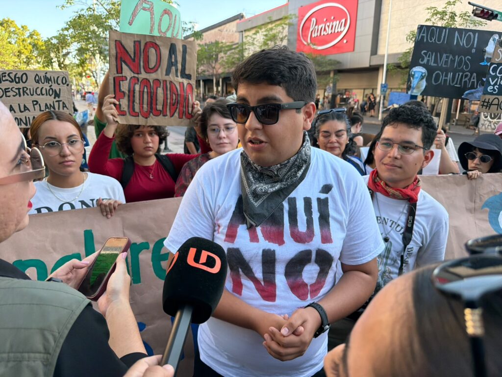 Hombre con una playera de Aquí No y personas detrás con pancartas