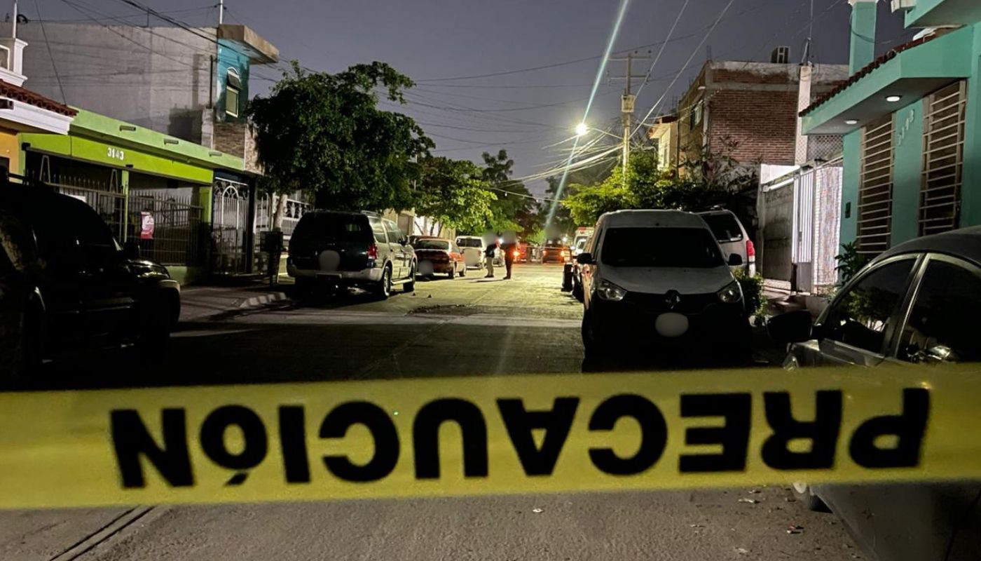 Cinta amarilla de precaución en una calle con carros estacionados y una unidad poliicaca al fondo