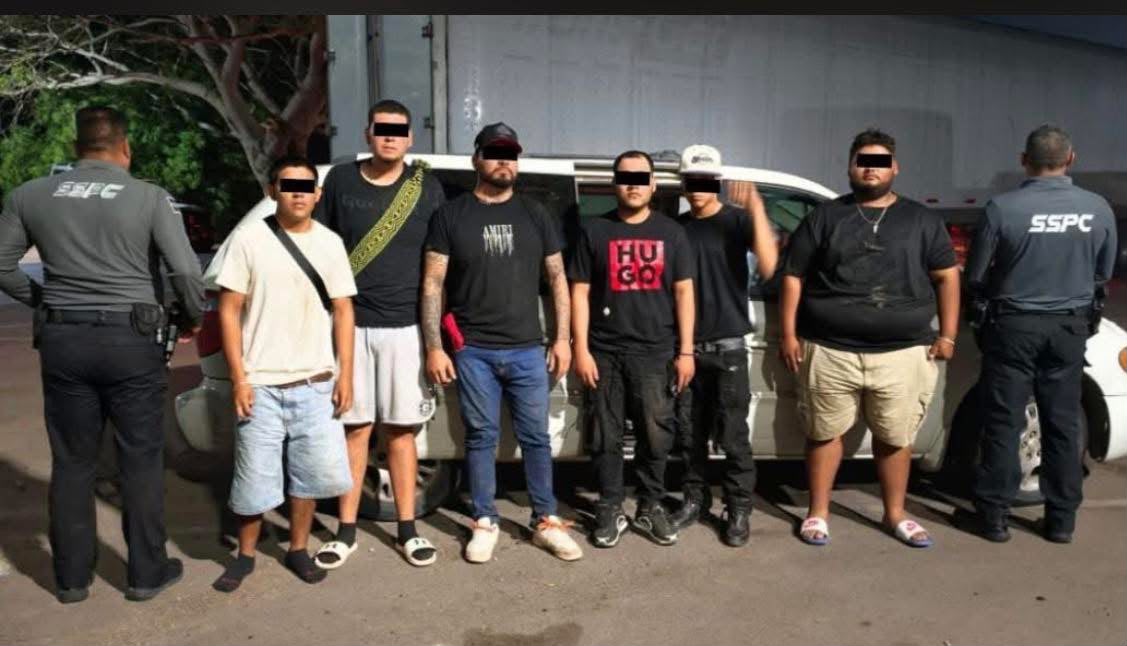 Seis personas con el rostro cubierto frente a una camioneta y a los costados, dos agentes de seguridad de espaldas
