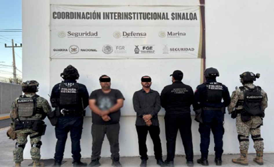 Agentes policiacos de espaldas y en el centro dos personas con el rostro cubierto y las manos esposadas
