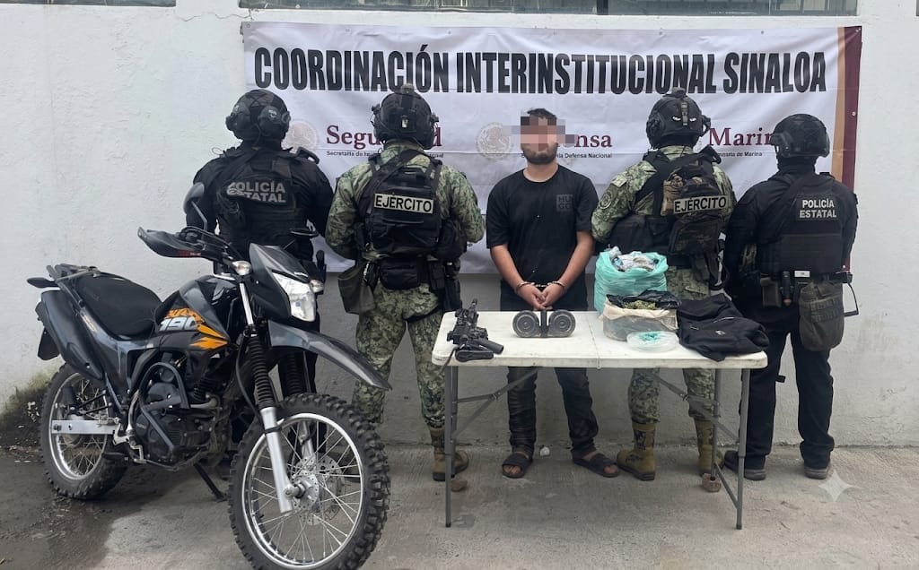 Elementos policicos de espaldas y un civil con el rostro cubierto y las manos esposadas al centro, una lona la fondo con logos y una mesa blanca con objetos, a un lado una motocicleta negra