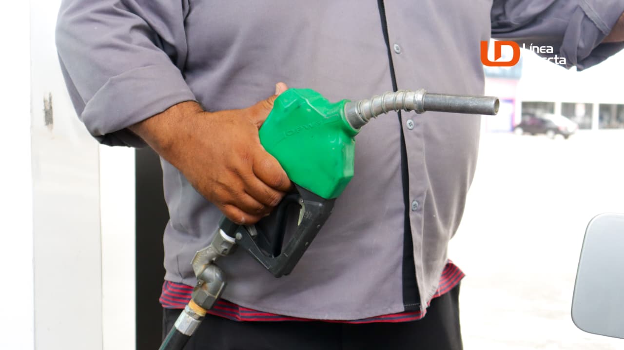 Mano sosteniendo una manguera dispensadora de gasolina de color verde y al fondo parte de una bomba de gasolina