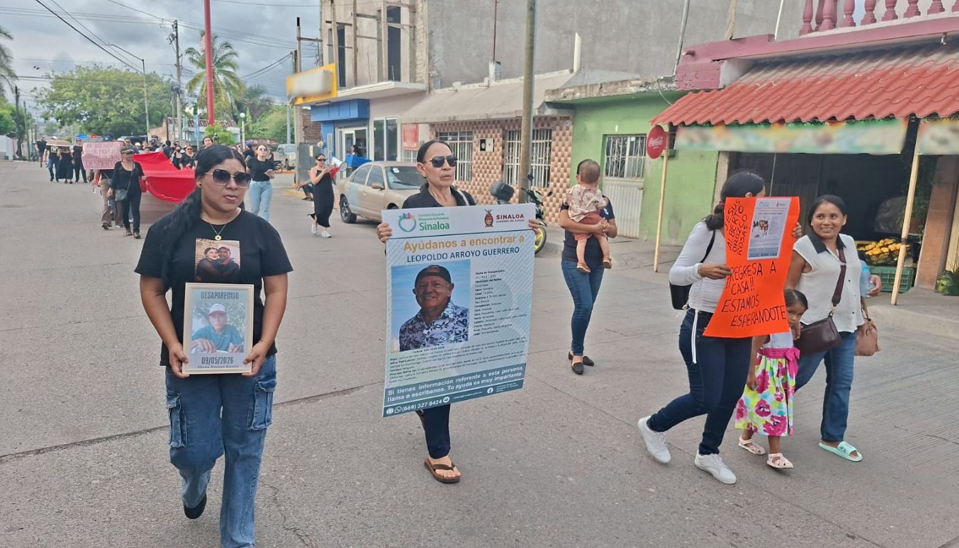 Personas realizando una manifestación con pancartas y fichas de búsqueda, mientras caminan por una calle con carros y negocios