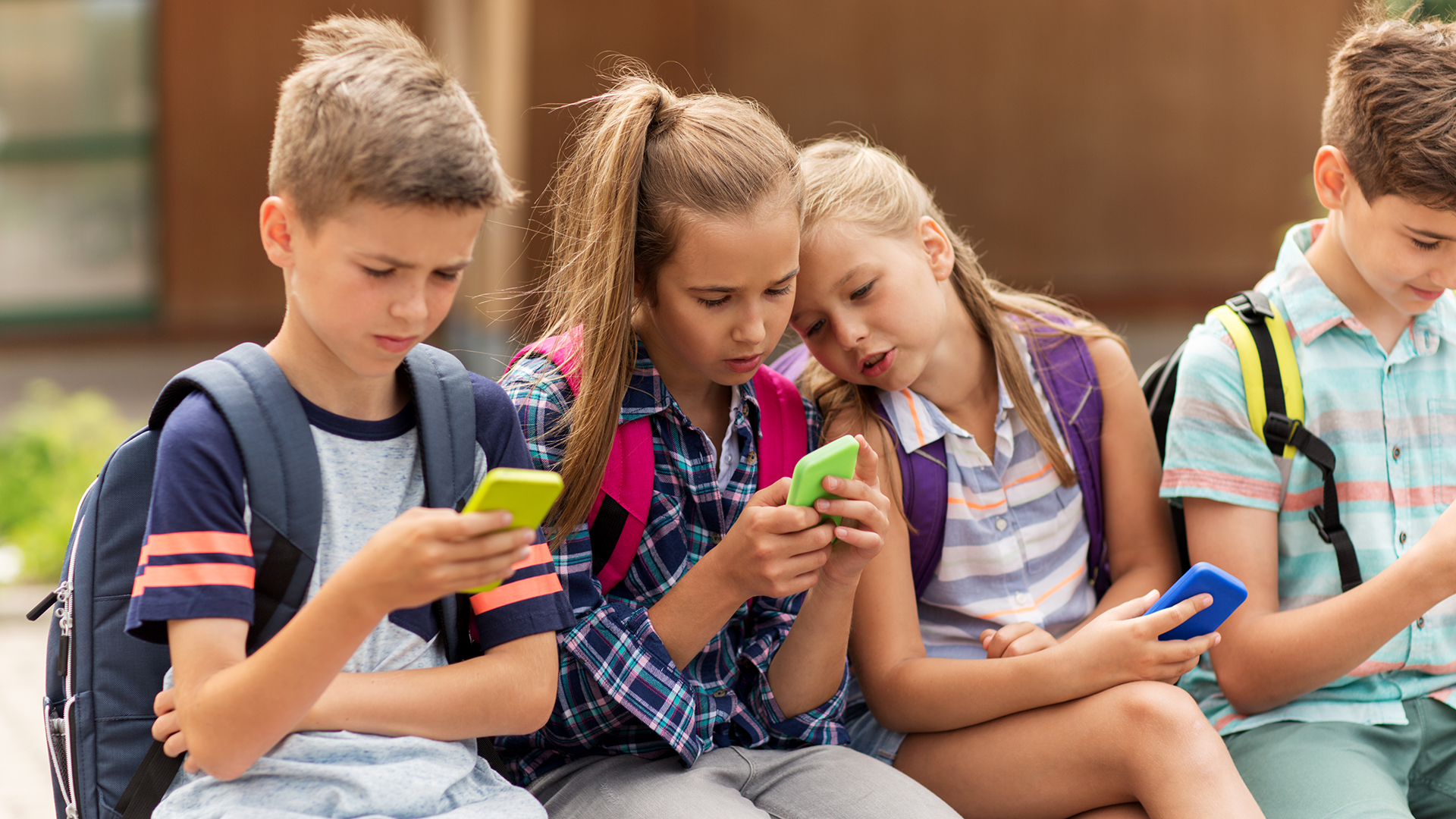 Niños con mochilas y celulares sentados en una jardinera