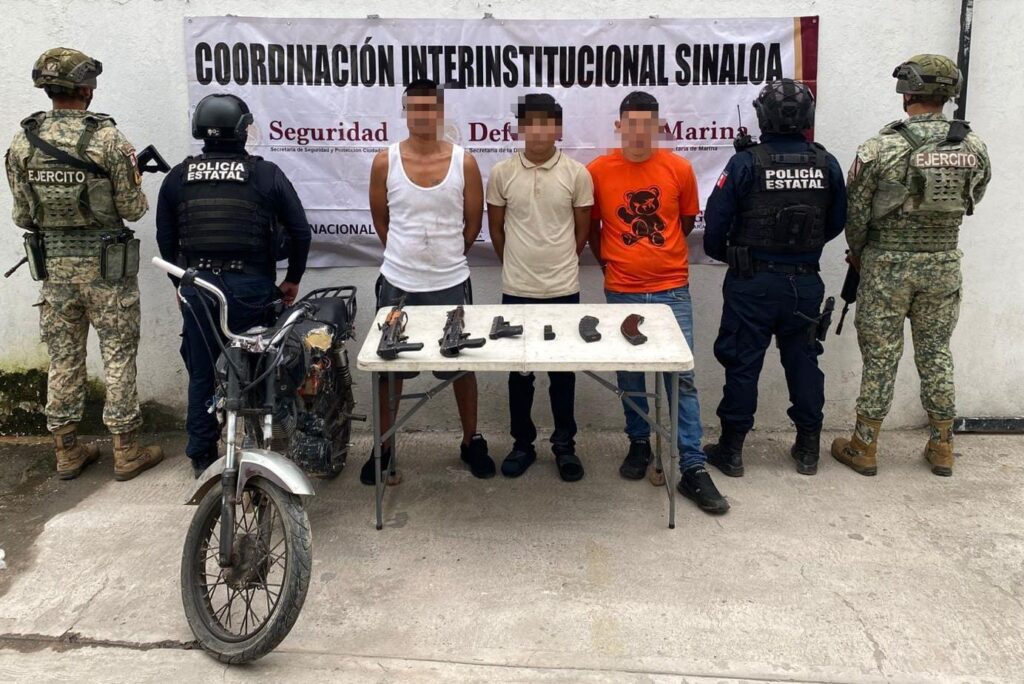 Elementos policiacos de espaldas y tres civiles con el rostro cubierto y las manos esposadas, una mesa con objetos y una motocicleta